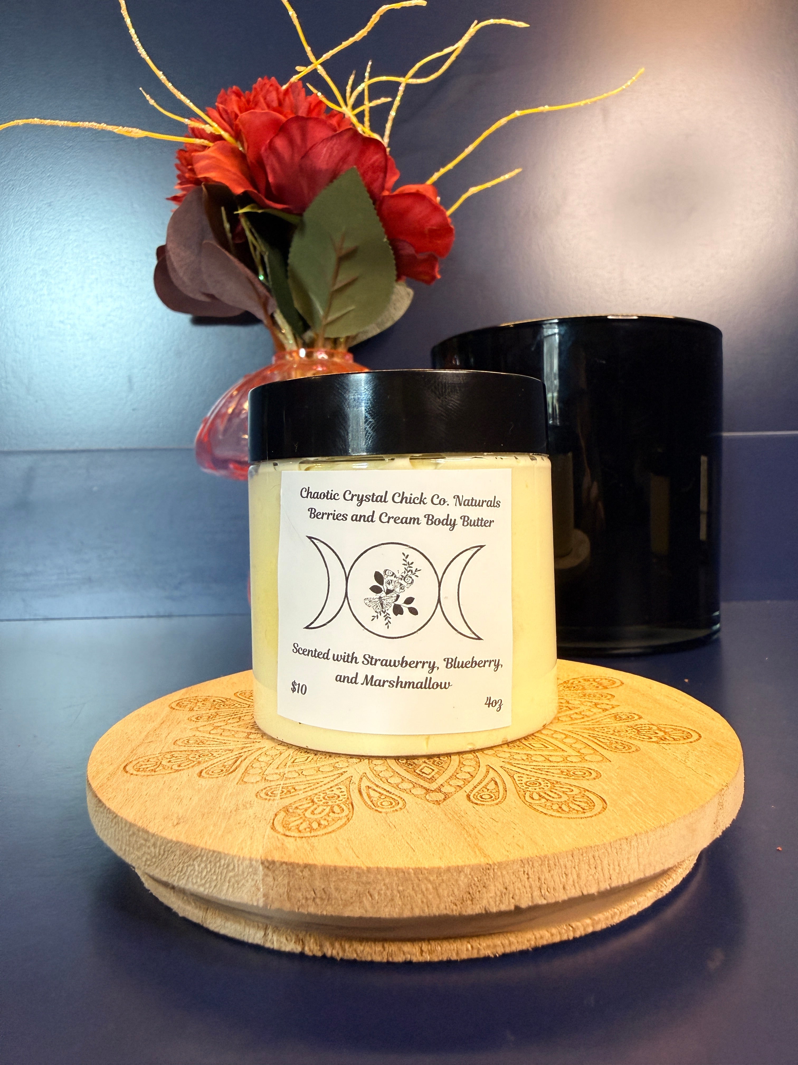 Jar of yellow body butter with a label on a decorative wooden coaster in front of a red flower and black jar