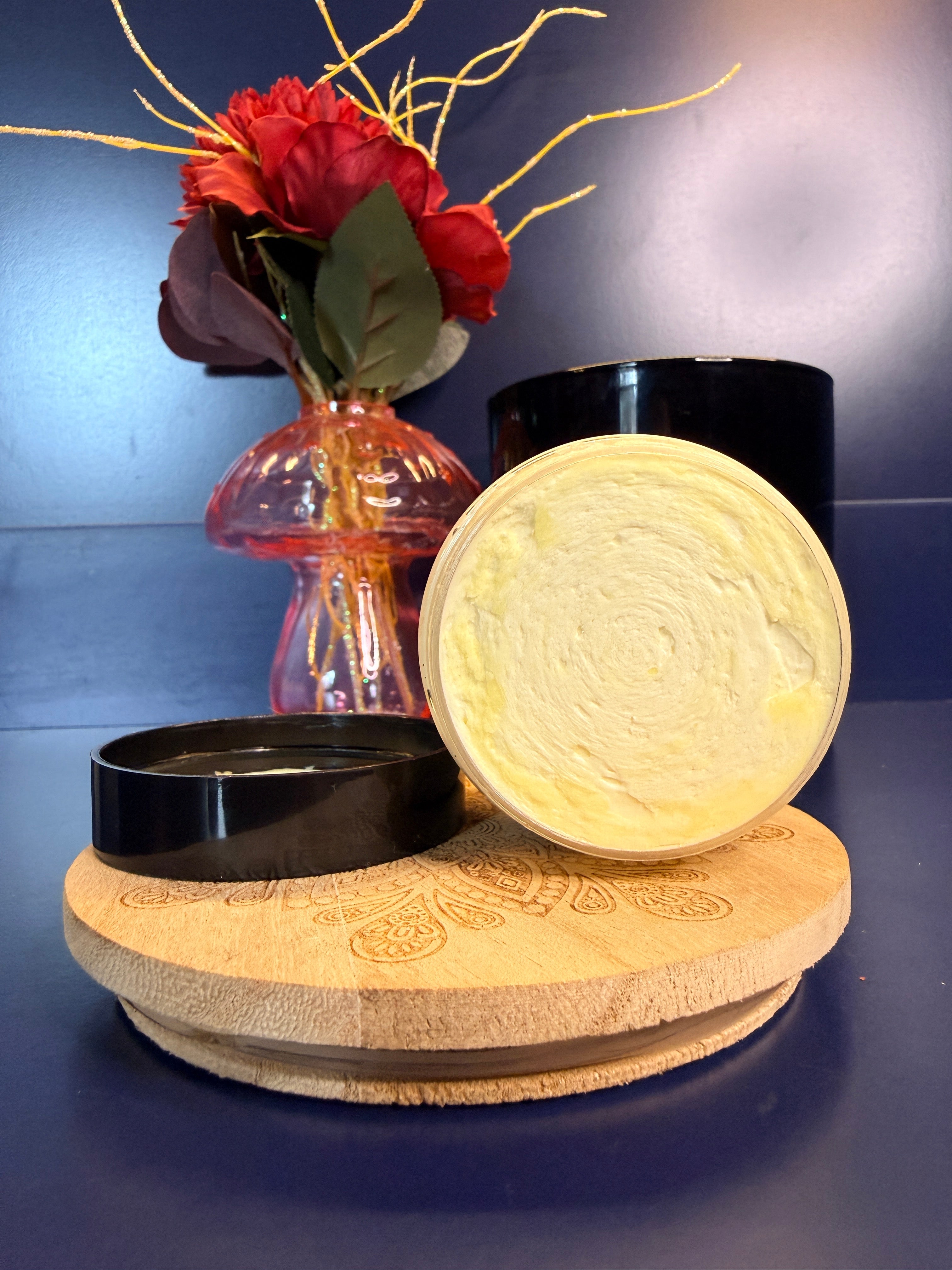 Open jar of yellow body butter on a decorative wooden coaster in front of a red flower and black jar
